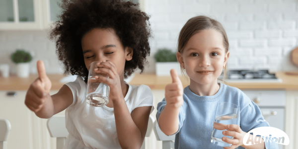 Ein Mädchen mit dunkler Haut und einem Afro und ein Mädchen mit weißer Haut und einem Zopf sind in einer Küche mit hellen Bauernmöbel. Das Mädchen mit weißer Haut schaut in die Kamera und hält in der einen Hand ein Wasserglas und mit der anderen Hand den Daumen nach oben. Das andere Mädchen ist damit beschäftig aus dem Glas zu trinken und hält auch den Daumen nach oben.