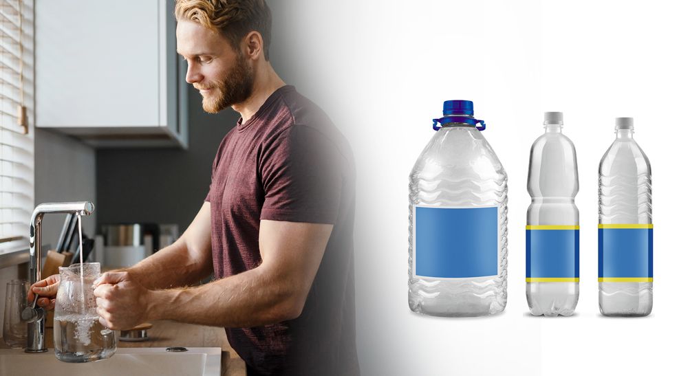 Mann steht an einem Wasserhahn in der Küche und füllt Leitungswasser in eine Glaskaraffe. Daneben stehen drei Mineralwasserflaschen im Bild.