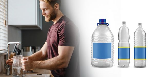 Mann steht an einem Wasserhahn in der Küche und füllt Leitungswasser in eine Glaskaraffe. Daneben stehen drei Mineralwasserflaschen im Bild.