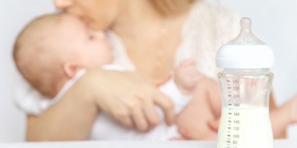 Eine blonde Frau hält ein Baby im Arm und küsst es. Im Vordergrund des Bildes steht eine Babyflasche, die zur Hälfte mit flüssiger Babynahrung gefüllt ist.