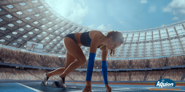 Eine Läuferin in einem blauen Laufset ist in einem voll besetzten Sportstadion und befindet sich in der Startposition, um mit dem Rennen zu beginnen. Sie hat sich auf ihren Fingern abgestützt und die Füße auf die Starthilfe gestellt.