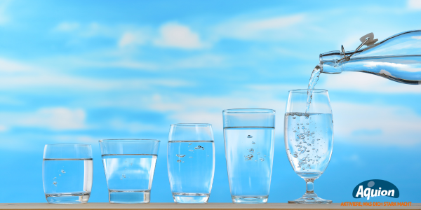 Auf einem Brett stehen 5 unterschiedlich große Gläser von schlicht zu schön, von klein zu groß. In das letzte Glas wird aus einer Glasflasche Trinkwasser geschüttet.Im Hintergrund ein blauer Himmel mit Schleier